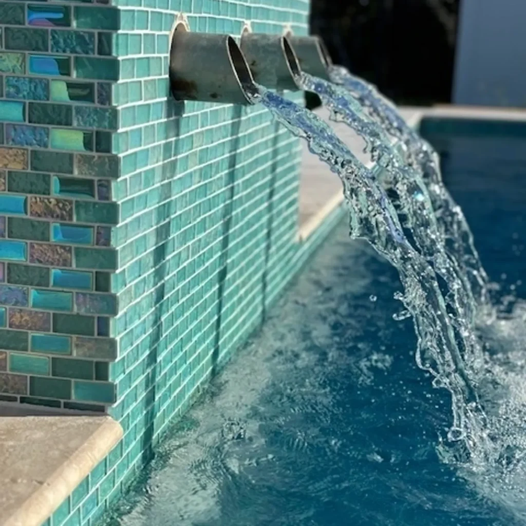 Close-up of three bronze scupper spouts pouring water over an iridescent green glass tile wall into a pool.
