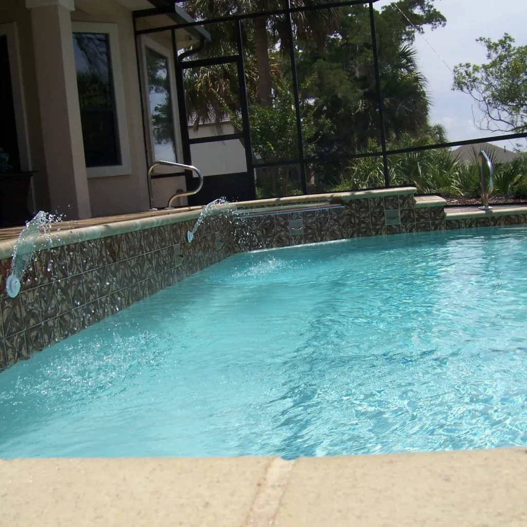 A screened rectangular backyard pool with two deck jets spraying water over a tiled raised wall.