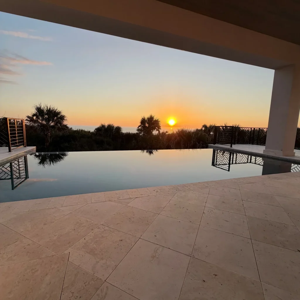An infinity-edge pool overlooks the ocean at sunset from a covered terrace.