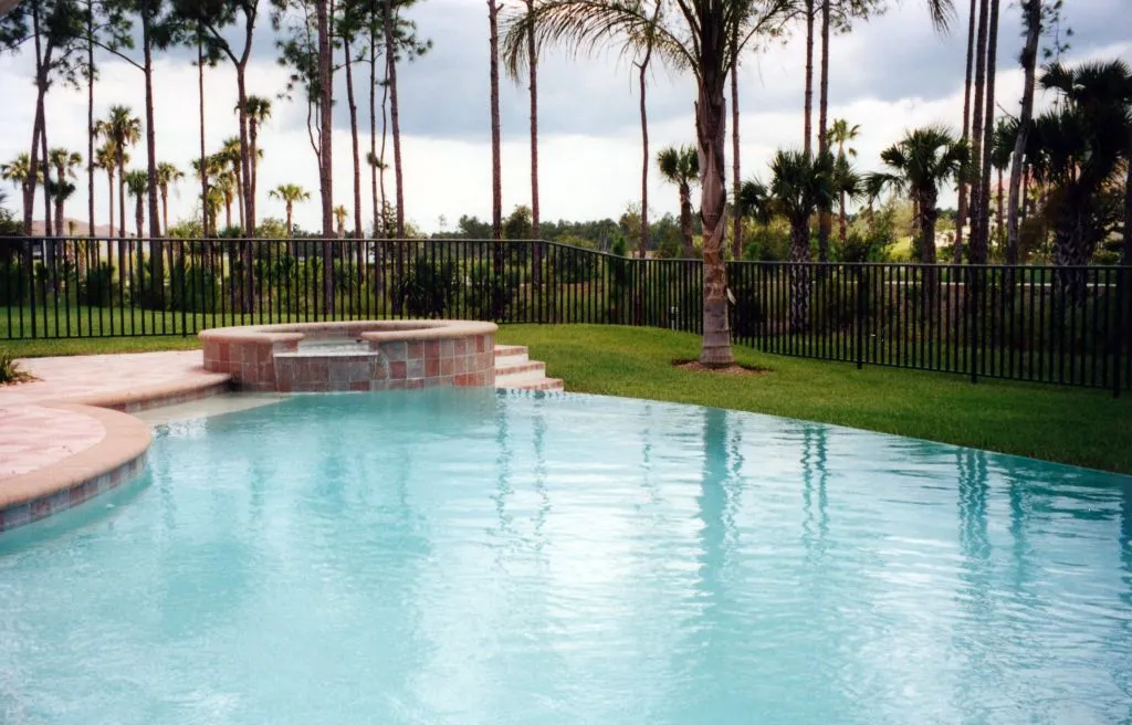 A backyard pool with a raised spa and steps, bordered by a metal fence and palm trees under an overcast sky.