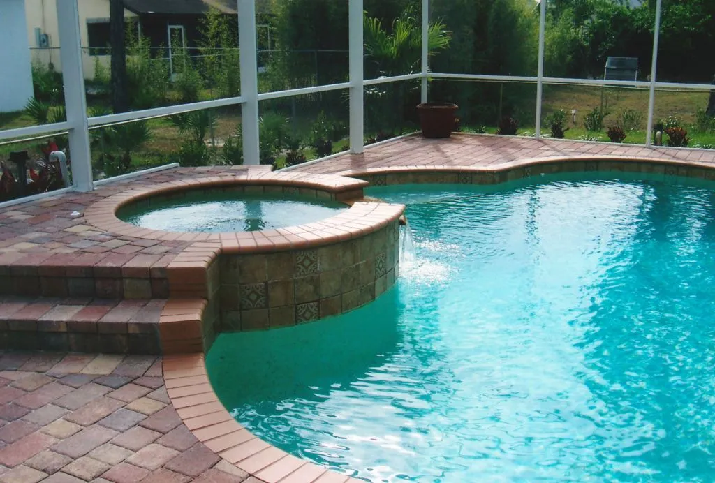 A raised round spa with tiled facing spills water into a freeform pool under a screened patio enclosure.