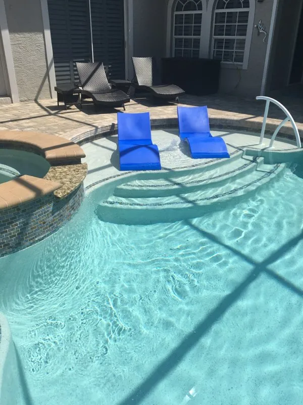 Two blue in-pool loungers sit in shallow water near a raised spa under a screened patio.
