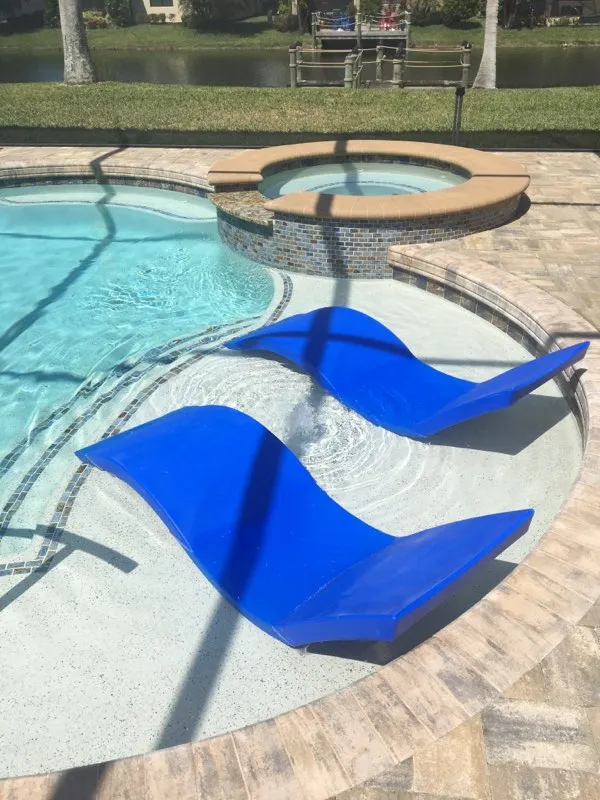 Two blue in-pool loungers sit on a sun shelf next to a raised round spa, with a canal visible beyond the screened lanai.