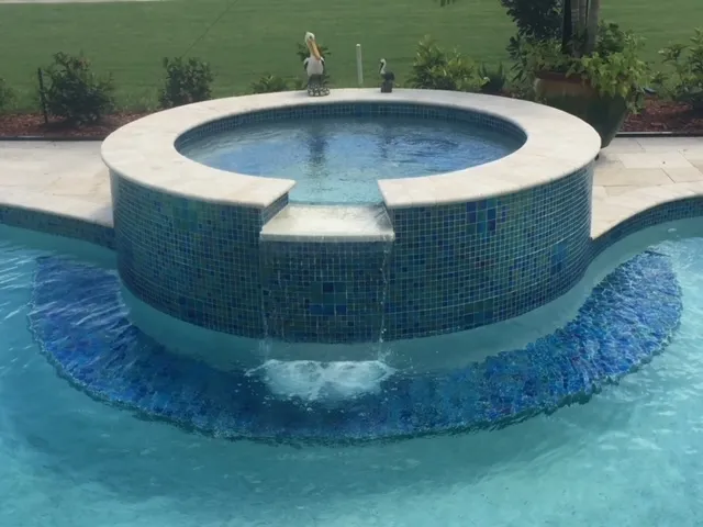 A round raised spa with blue mosaic tile spills water over its edge into the pool below.