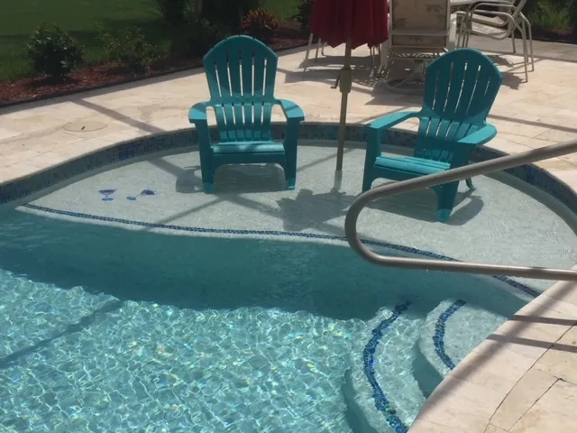 Two teal chairs sit on a submerged sun shelf at the edge of a pool with travertine decking.