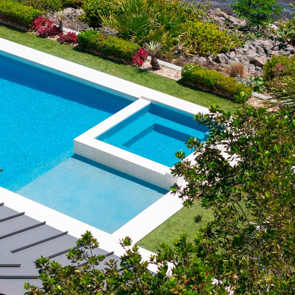 Aerial view of a modern rectangular pool with an attached raised spa and sun shelf next to a rocky waterfront.