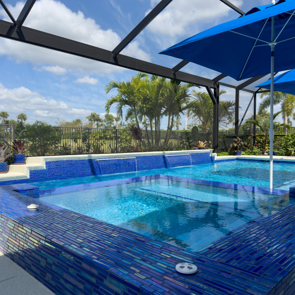 A raised spa with sheer descent waterfalls flows into a rectangular pool, both finished in vivid blue glass mosaic tile, under a screened lanai.