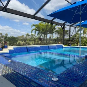 A raised spa with sheer descent waterfalls flows into a rectangular pool, both finished in vivid blue glass mosaic tile, under a screened lanai.