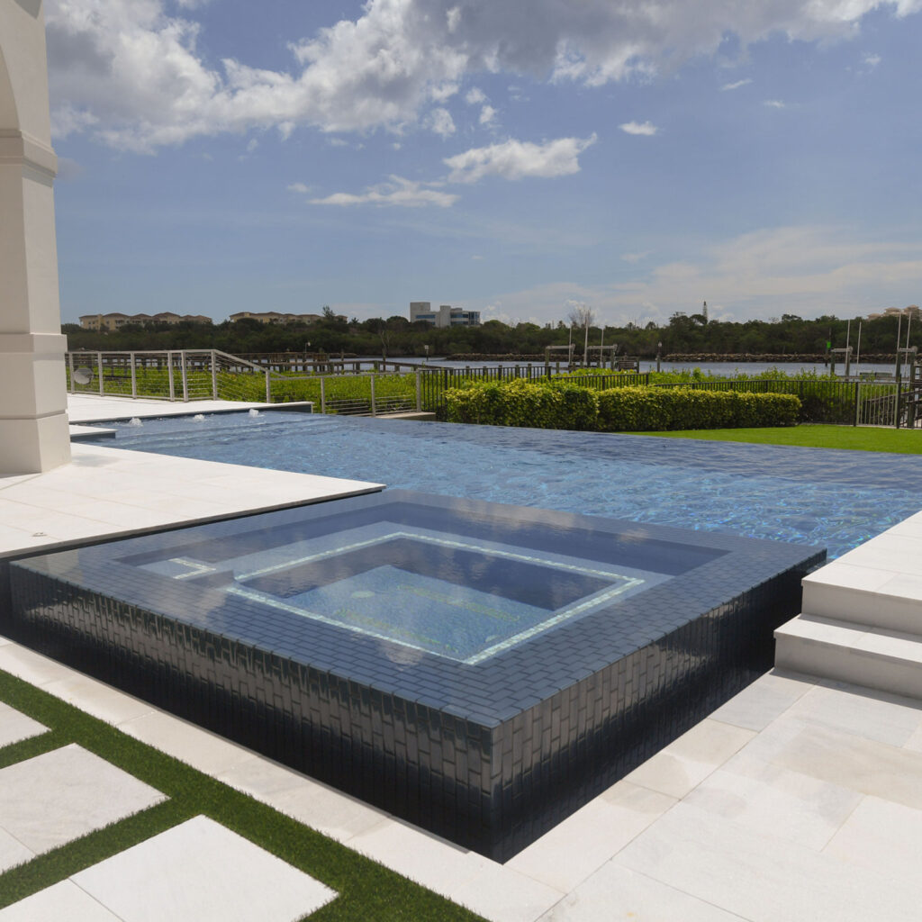 A raised glass-tiled spa sits in the foreground of an infinity-edge pool overlooking a waterway.