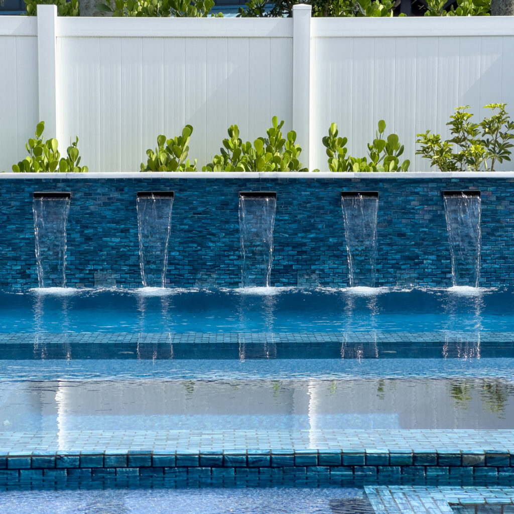 Five tiled water scuppers cascade into a blue pool in front of a white privacy fence.