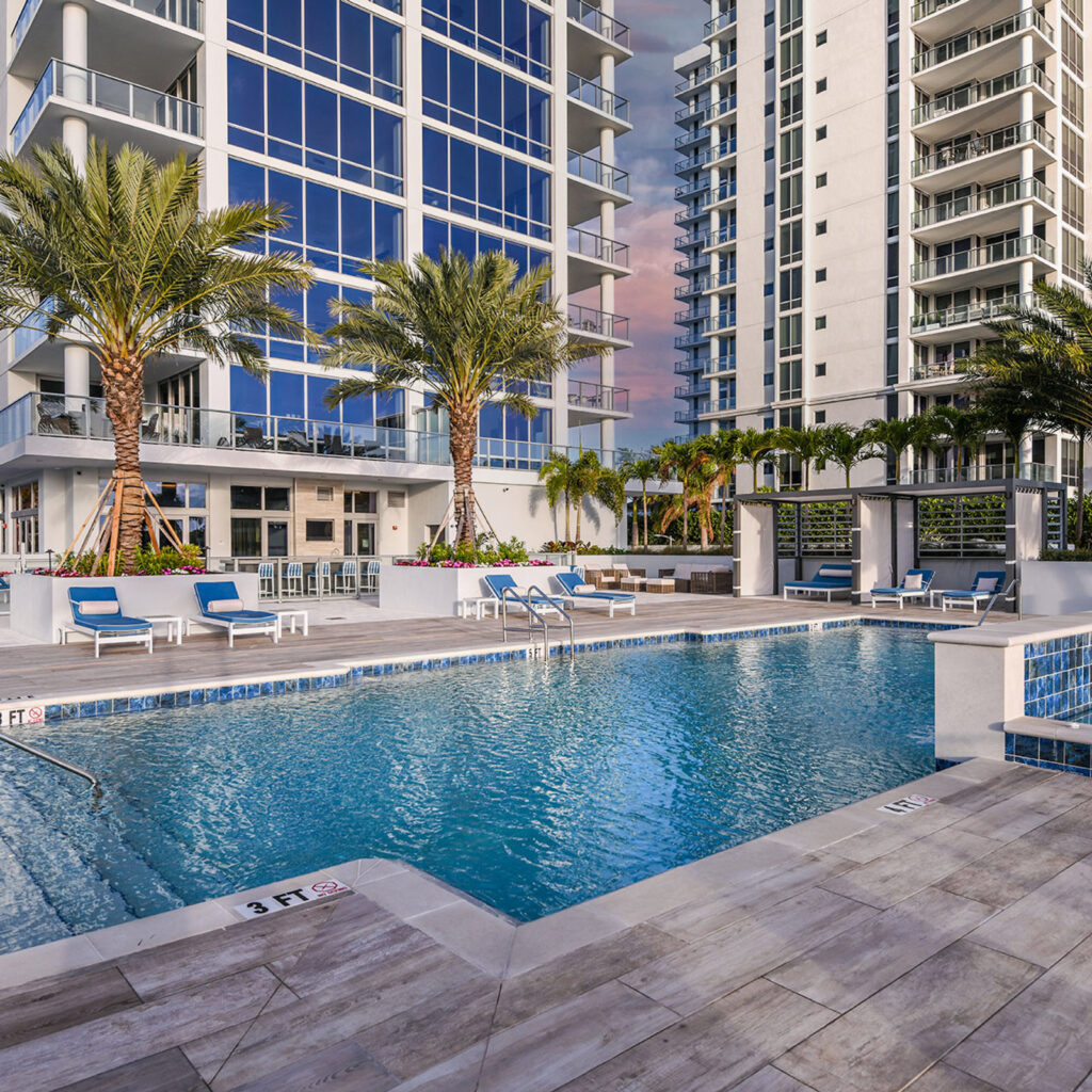 A rectangular pool at a high-rise residential tower amenity deck with lounge chairs and palm trees at dusk.