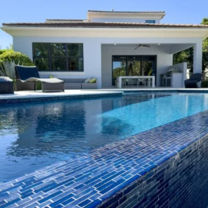 A rectangular infinity-edge pool with blue glass tile trim in front of a modern white house with covered patio.