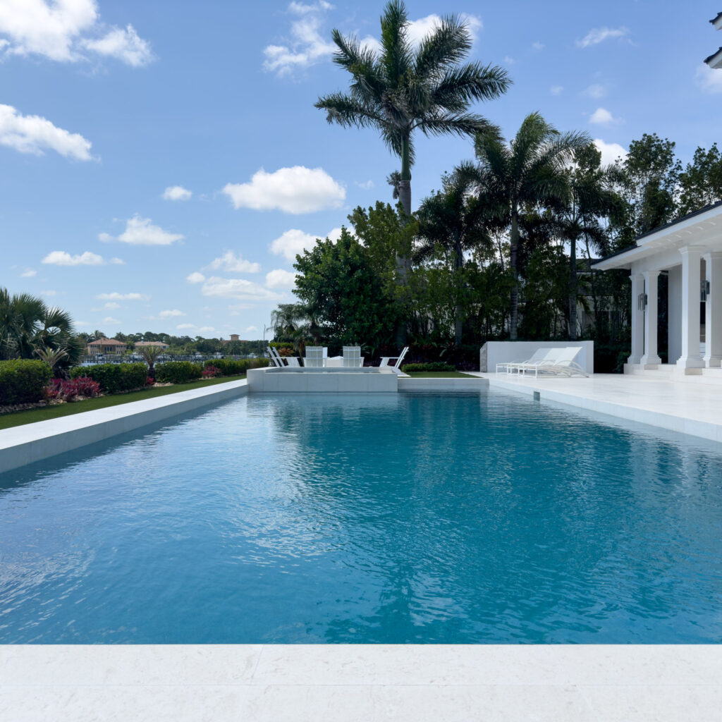 A long rectangular infinity-edge style pool with an integrated raised spa, flanked by white loungers and palm trees, with a canal visible in the distance.