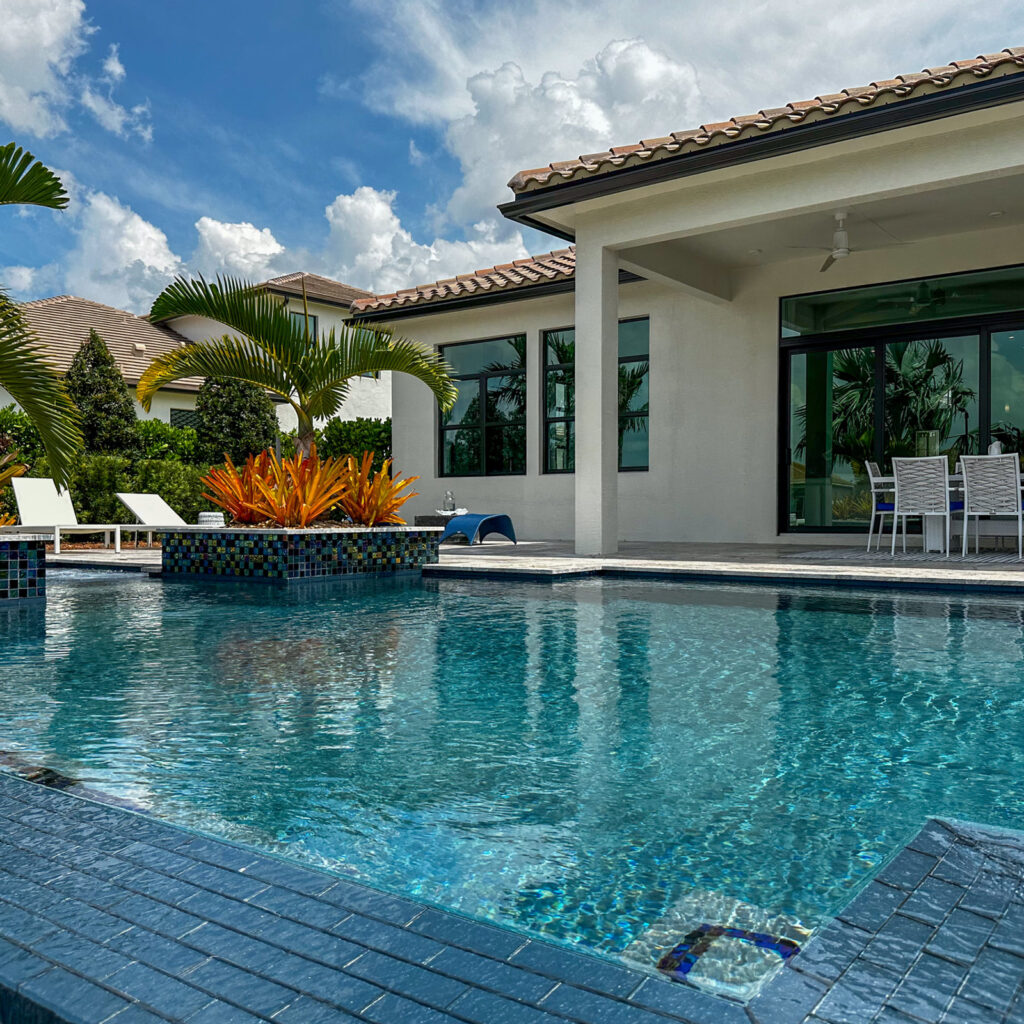 A freeform pool with a mosaic-tiled planter island reflects a modern white house under a blue sky.