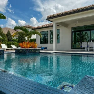A freeform pool with a mosaic-tiled planter island reflects a modern white house under a blue sky.