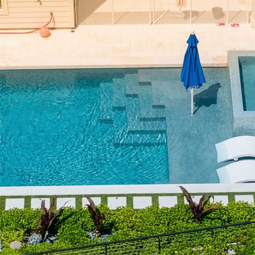 Aerial view of a pool with tiled entry steps, a closed blue umbrella, lounge chairs, and a raised spa in the corner.