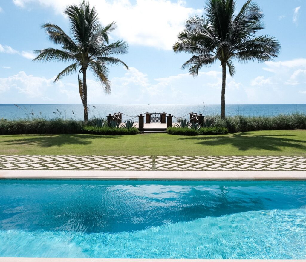A pool edge in the foreground with a lawn, palm trees, and ocean view in the background.