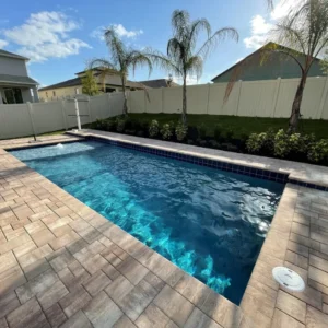 A small rectangular backyard pool with dark blue waterline tile and paver deck, surrounded by a vinyl privacy fence and palm trees.
