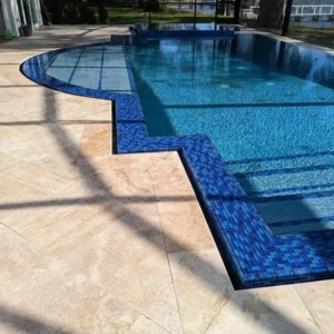 A freeform pool with blue mosaic tile edging and a broad tanning ledge, framed by travertine decking under a screen enclosure near the water.
