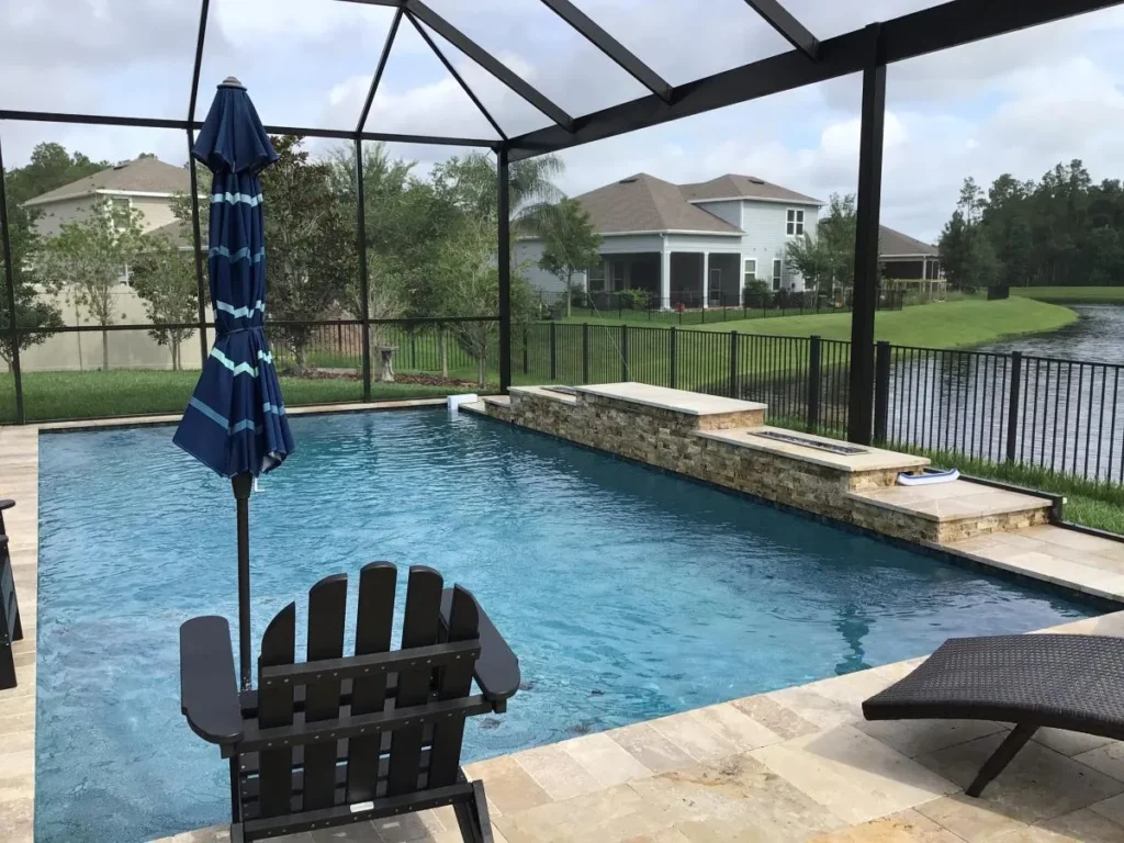 A screened rectangular pool with a raised stone fire feature beside a pond in a suburban backyard.