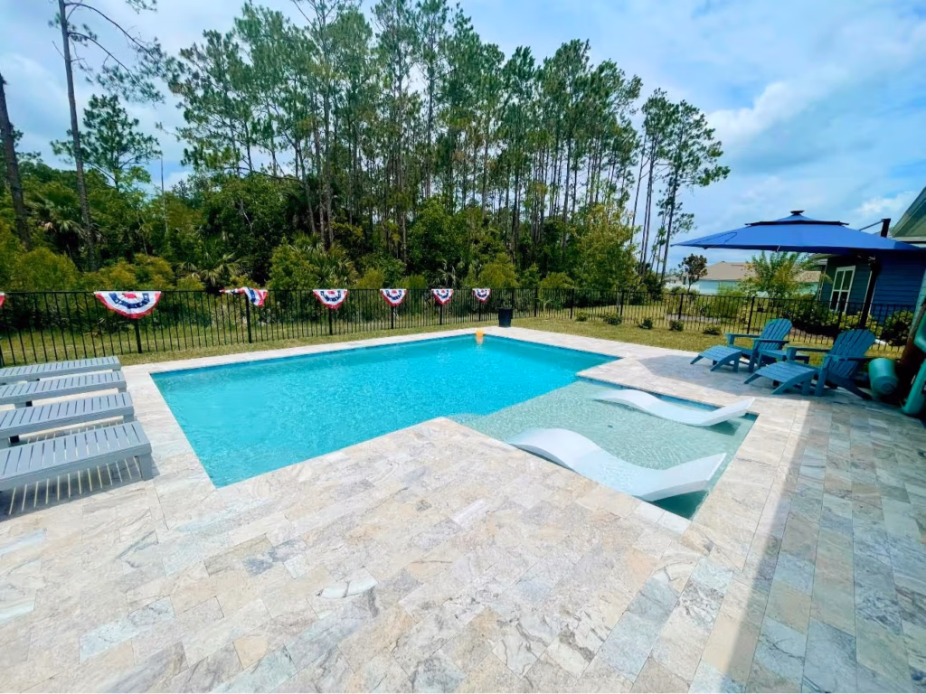An L-shaped backyard pool with a tanning ledge holding two in-pool loungers, surrounded by a travertine patio and pine trees.