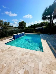 A rectangular backyard pool with a three-spout waterfall wall and travertine paver deck under a palm tree.