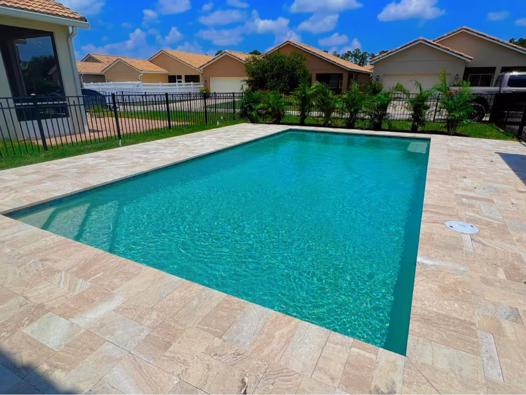 A rectangular pool with turquoise water and travertine deck in a suburban backyard with houses visible beyond the fence.