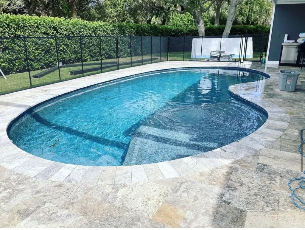 A kidney-shaped pool with turquoise water and travertine deck, enclosed by a black safety fence and hedges.