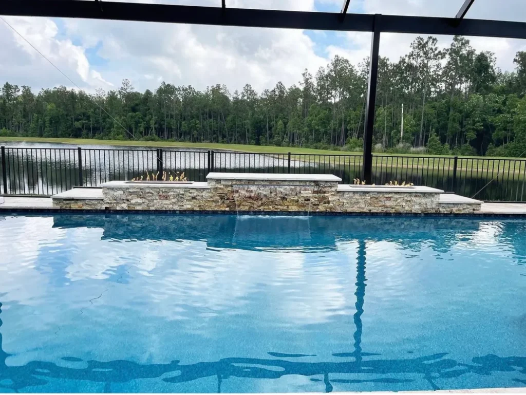 Rectangular pool with a raised stacked-stone wall featuring two fire bowls and waterfall spouts, overlooking a pond and screened enclosure.