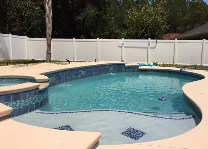 A freeform backyard pool with a raised spa and blue tile accents behind a white vinyl privacy fence.