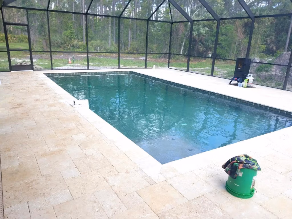 A newly built rectangular pool with travertine deck inside a screened enclosure, backed by trees and cleared land.