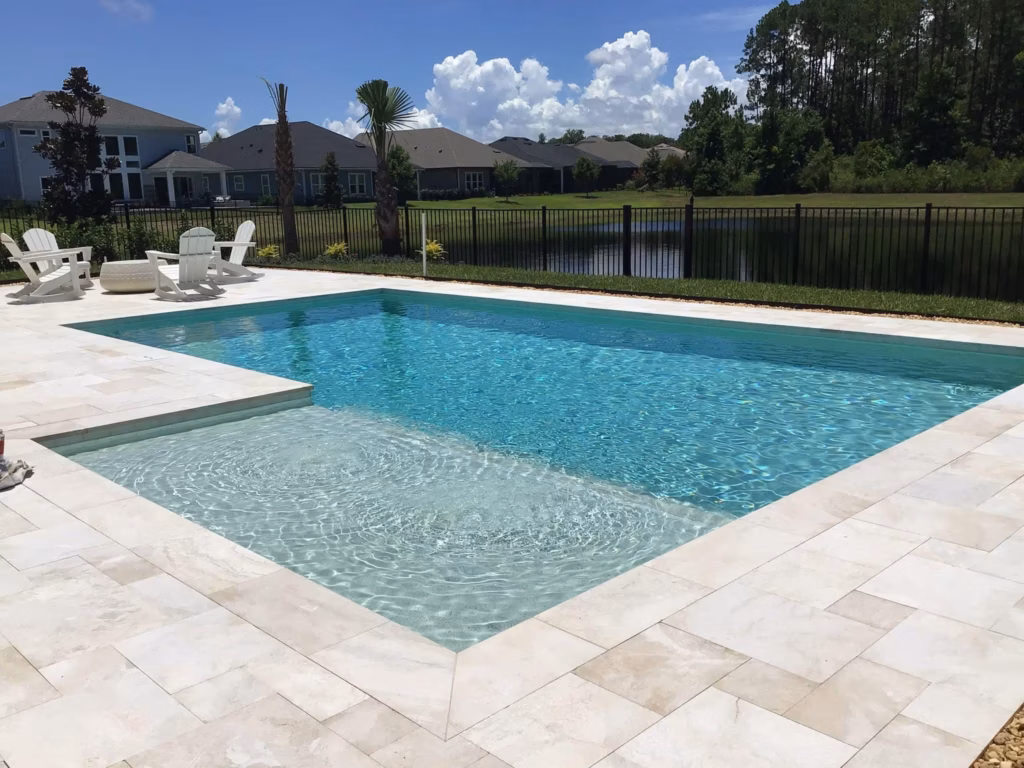 A rectangular pool with a shallow tanning ledge and travertine deck, overlooking a fenced pond in a suburban backyard.