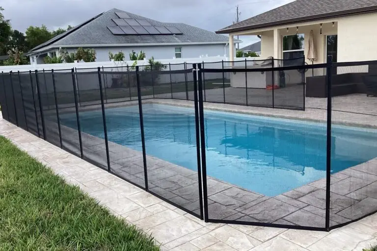 A rectangular backyard pool enclosed by a black mesh safety fence on a stamped concrete patio.