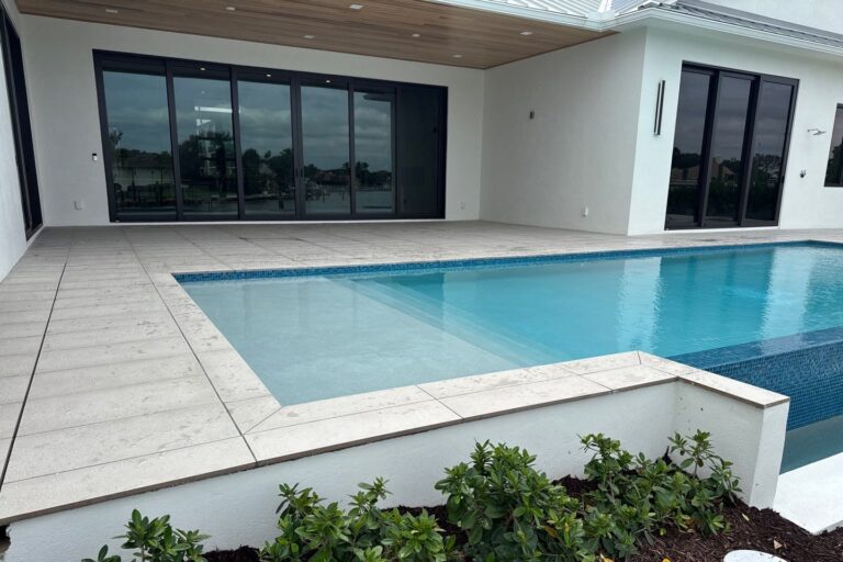 A modern rectangular pool with a tile waterline sits along a sleek white house with large glass sliding doors.