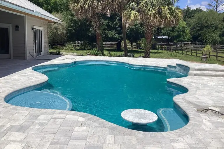 A freeform pool with a paver deck, palm trees, and a rural fenced pasture in the background.