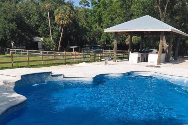 A freeform pool with a raised spa and waterfall feature next to a pavilion outdoor kitchen and horse pasture.