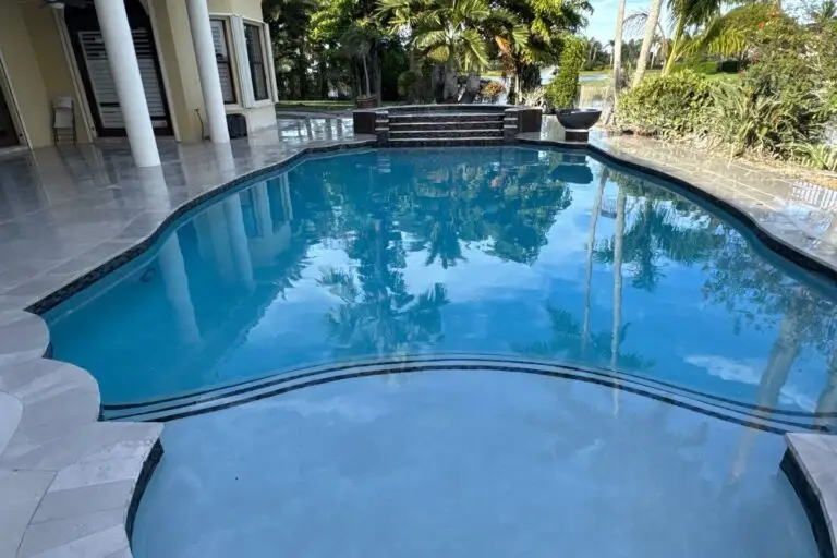 A large freeform swimming pool with a tiled waterline sits on a travertine patio behind a house, with a small water feature and steps at the far end.