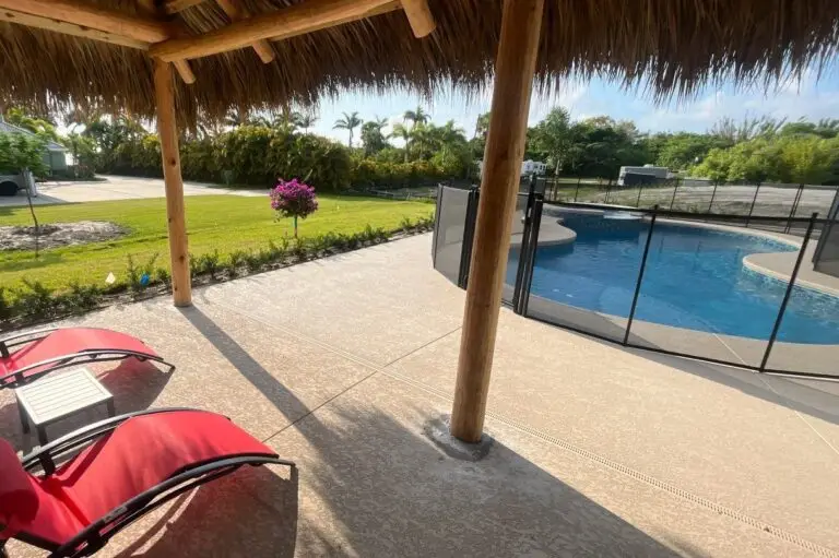 View of a fenced backyard pool seen from beneath a thatched-roof tiki hut with red loungers.