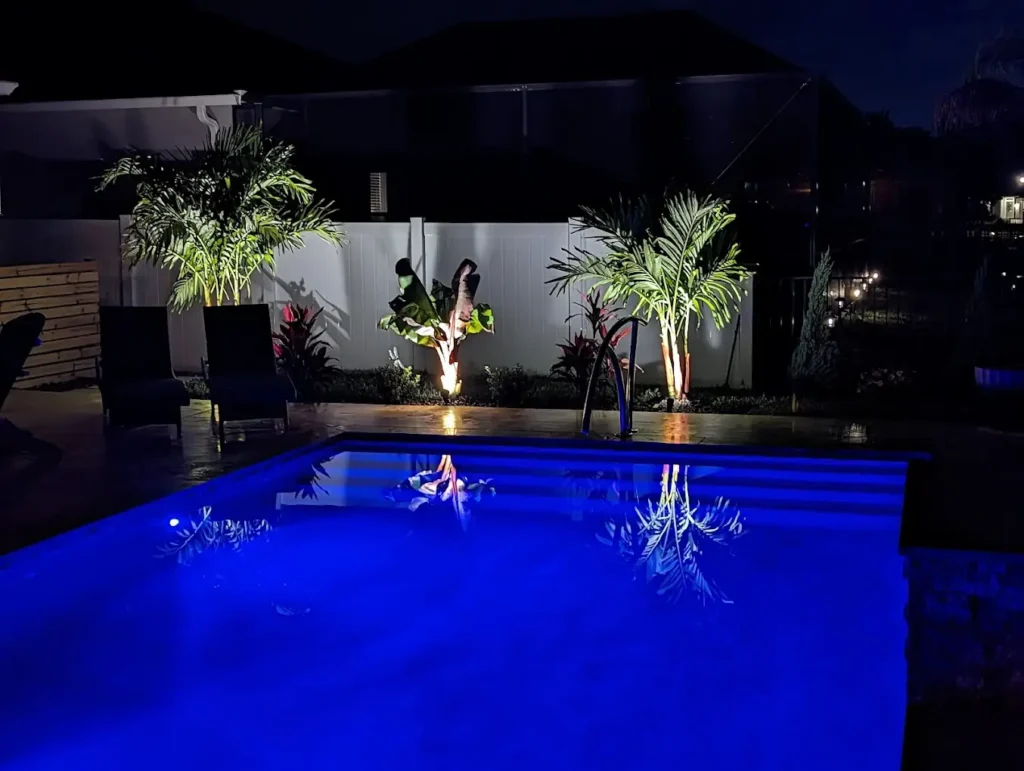 A rectangular backyard pool glowing blue at night surrounded by lit palm trees and a white vinyl fence.