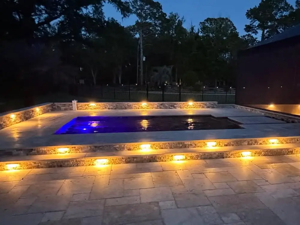 A small rectangular pool glowing blue at night on a raised travertine deck outlined with warm LED lighting.