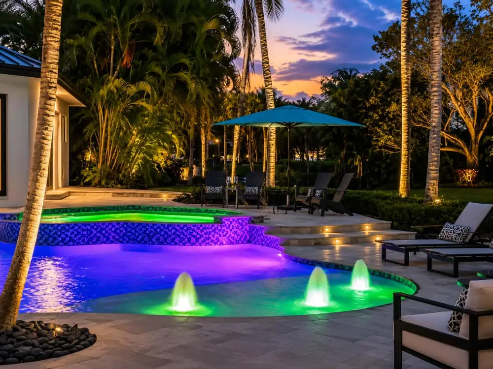 A freeform pool with a raised spa glows purple and green from LED lighting at dusk, surrounded by palm trees and a travertine patio.