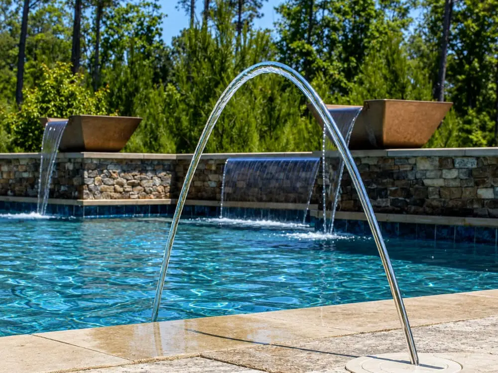 An arching water jet and stone scupper waterfalls cascade into a pool along a raised stone wall.