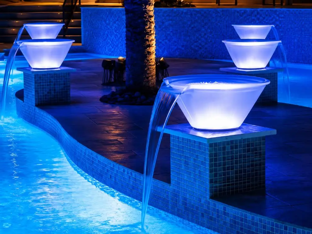 Illuminated white water bowls cascade into a glowing blue pool at night.