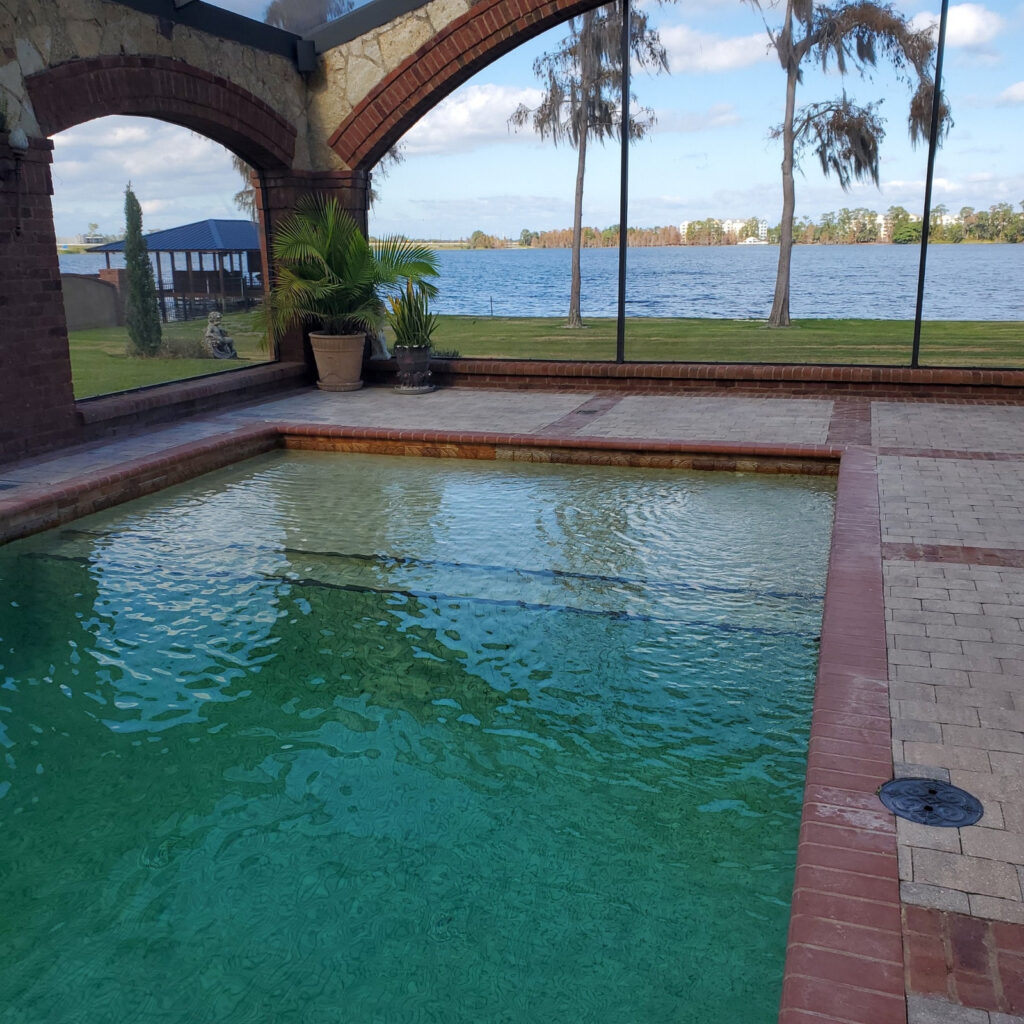 A rectangular pool with green-tinted water sits under a screened lanai overlooking a lake.