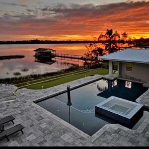 An infinity-edge style rectangular pool with a raised spa overlooks a lake at sunset, next to a putting green.