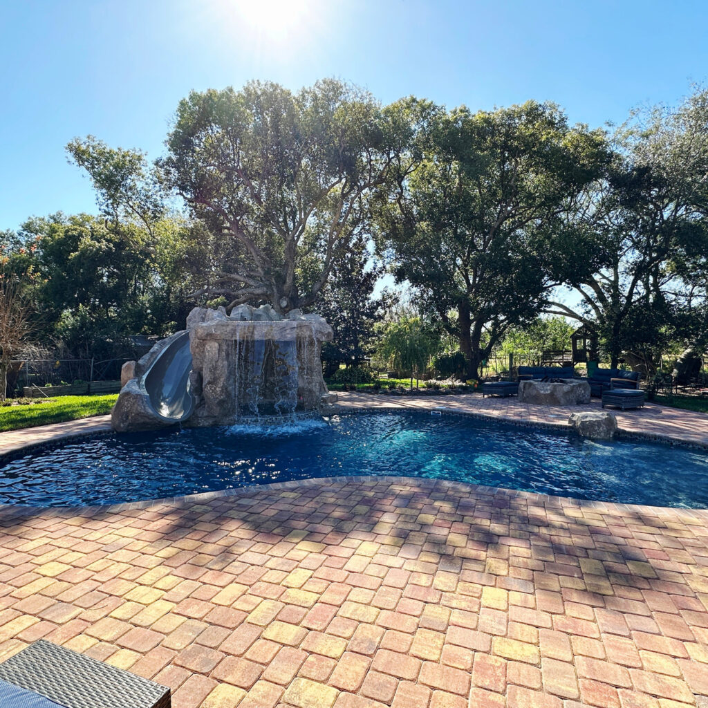 Freeform pool with a rock waterfall and slide surrounded by a paver patio and mature trees.
