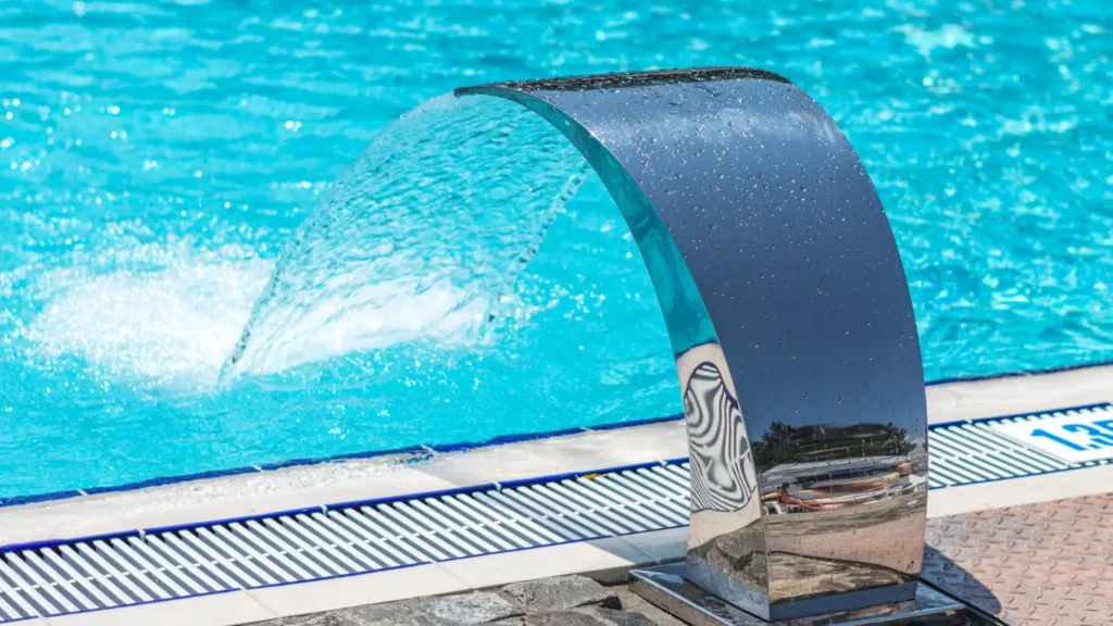 A stainless steel waterfall spout pours into a bright blue pool with a numbered lane marker on the tile edge.