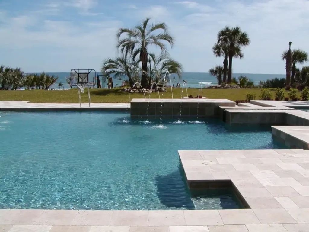 A large geometric pool with deck jets and a raised tile wall overlooks the ocean beyond a grassy yard.