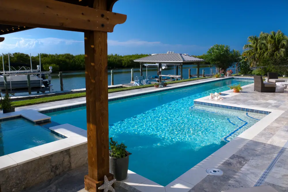 A long lap pool with travertine deck overlooking a canal with boats docked nearby.