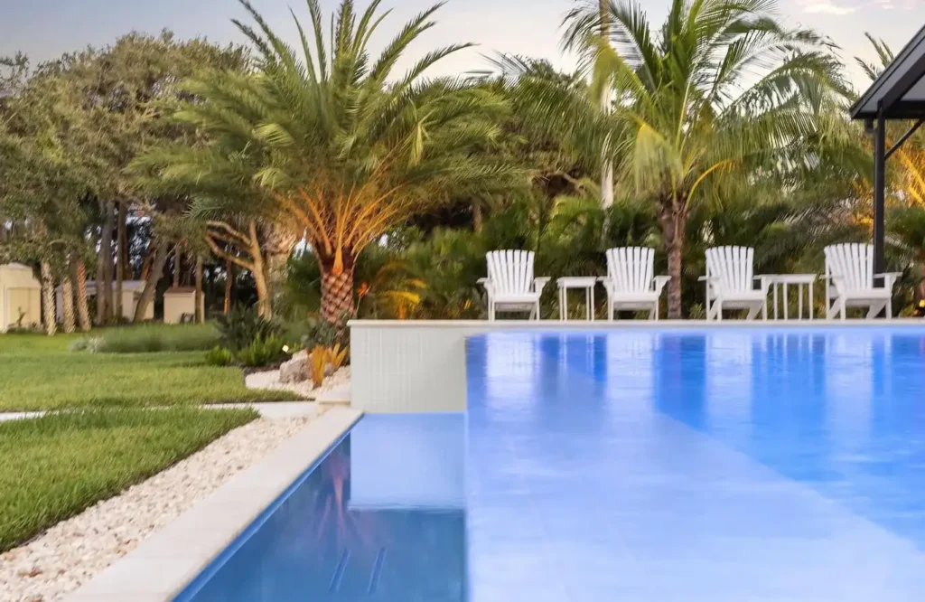 A pool edge with a tiled raised wall opens onto a landscaped backyard with palm trees and white lounge chairs at dusk.